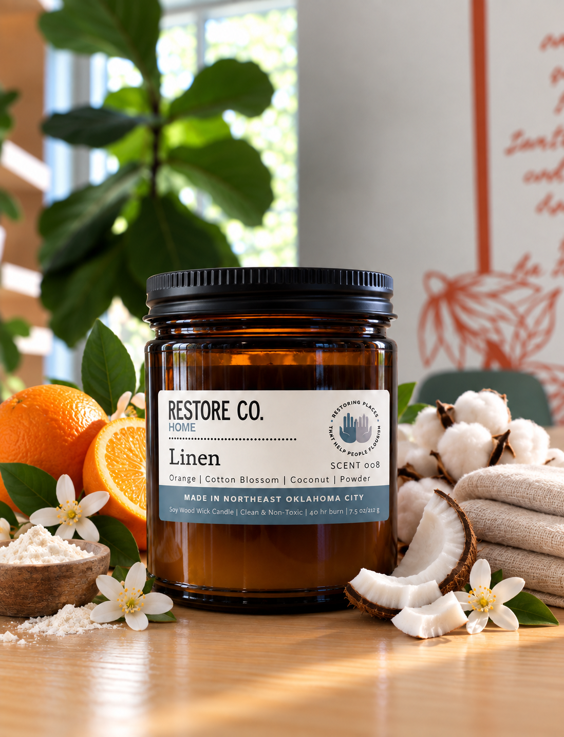 An amber glass jar candle with a black lid and a white label reading 'RESTORE CO. HOME' and 'Linen' sits on a wooden surface surrounded by orange slices, cotton bolls, flowers, and folded fabric in a bright, styled setting.