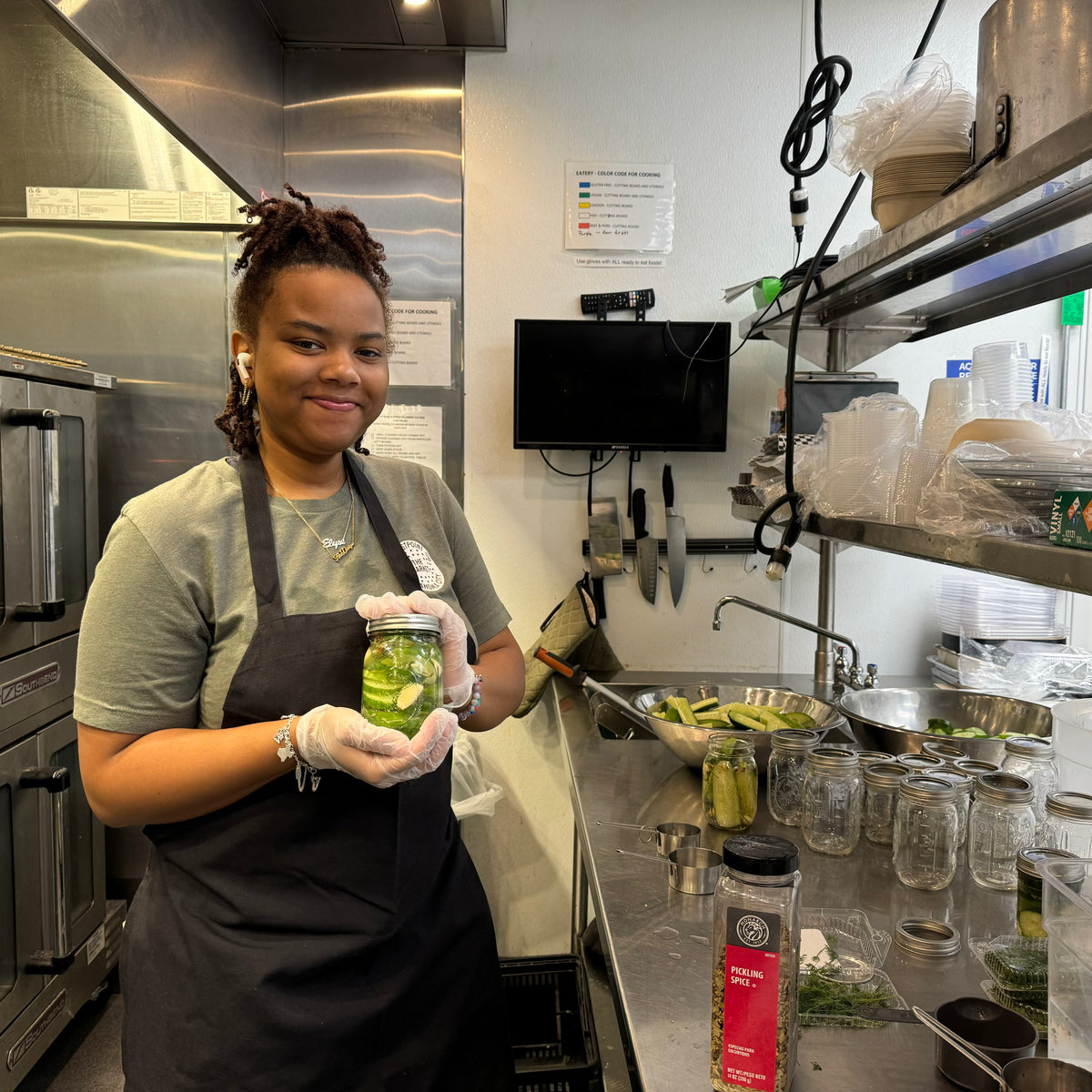 A woman wearing a black apron and white gloves holds a glass jar filled with pickled cucumbers in a commercial kitchen setting.