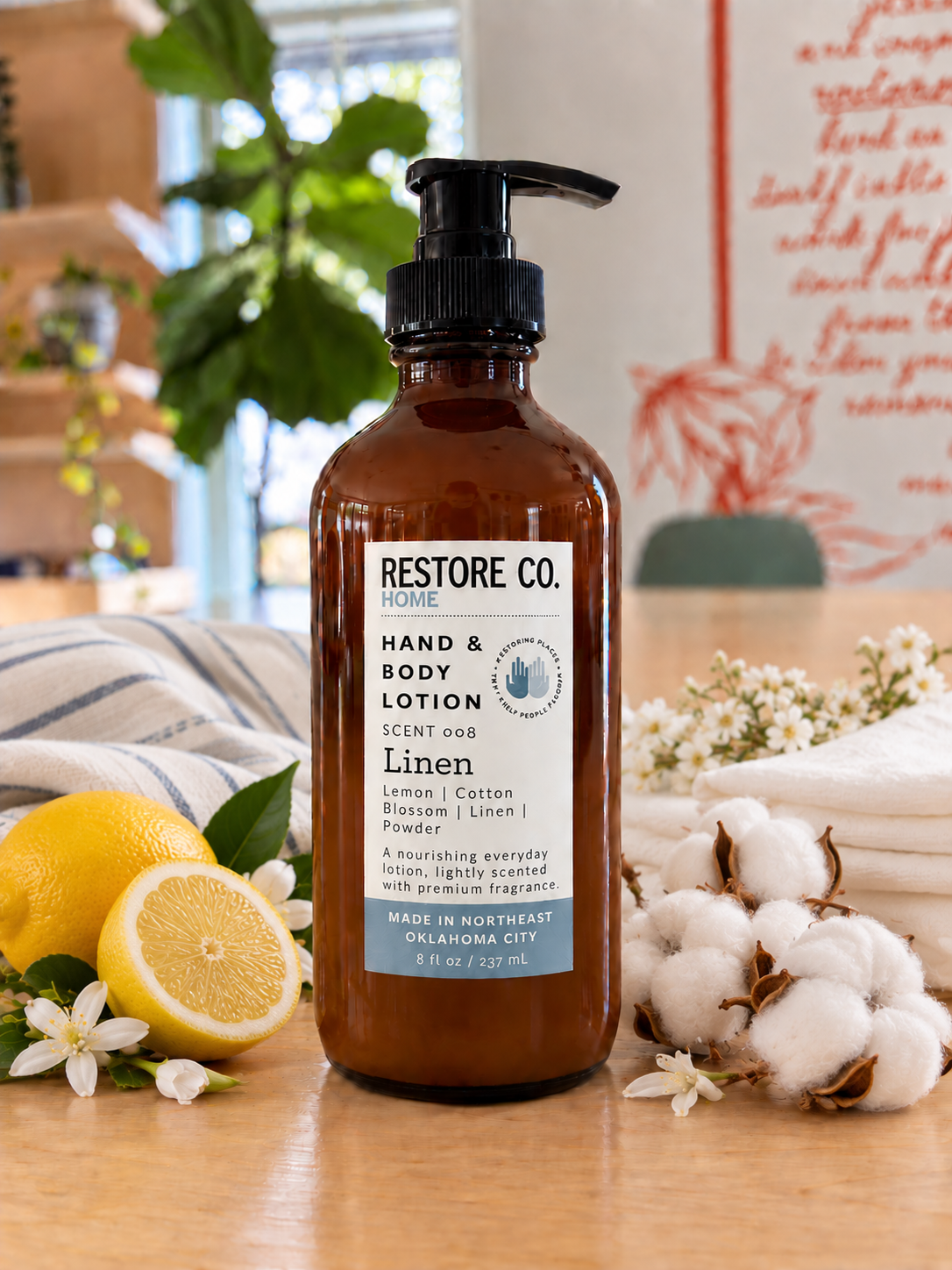 A bottle of Restore Co. Home Hand & Body Lotion in the Linen scent, styled on a wooden table with lemons, cotton, and folded towels in a bright, softly blurred indoor setting.