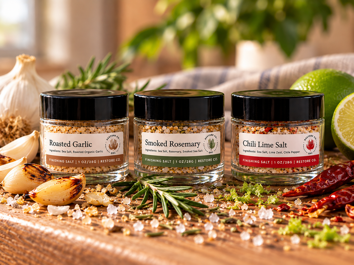 Three glass jars of seasoning salts are arranged on a wooden board with garlic, rosemary, chili peppers, and lime slices in a kitchen setting. The jars are labeled Roasted Garlic, Smoked Rosemary, and Chili Lime Salt, with the brand name Retime Co visible on the labels.