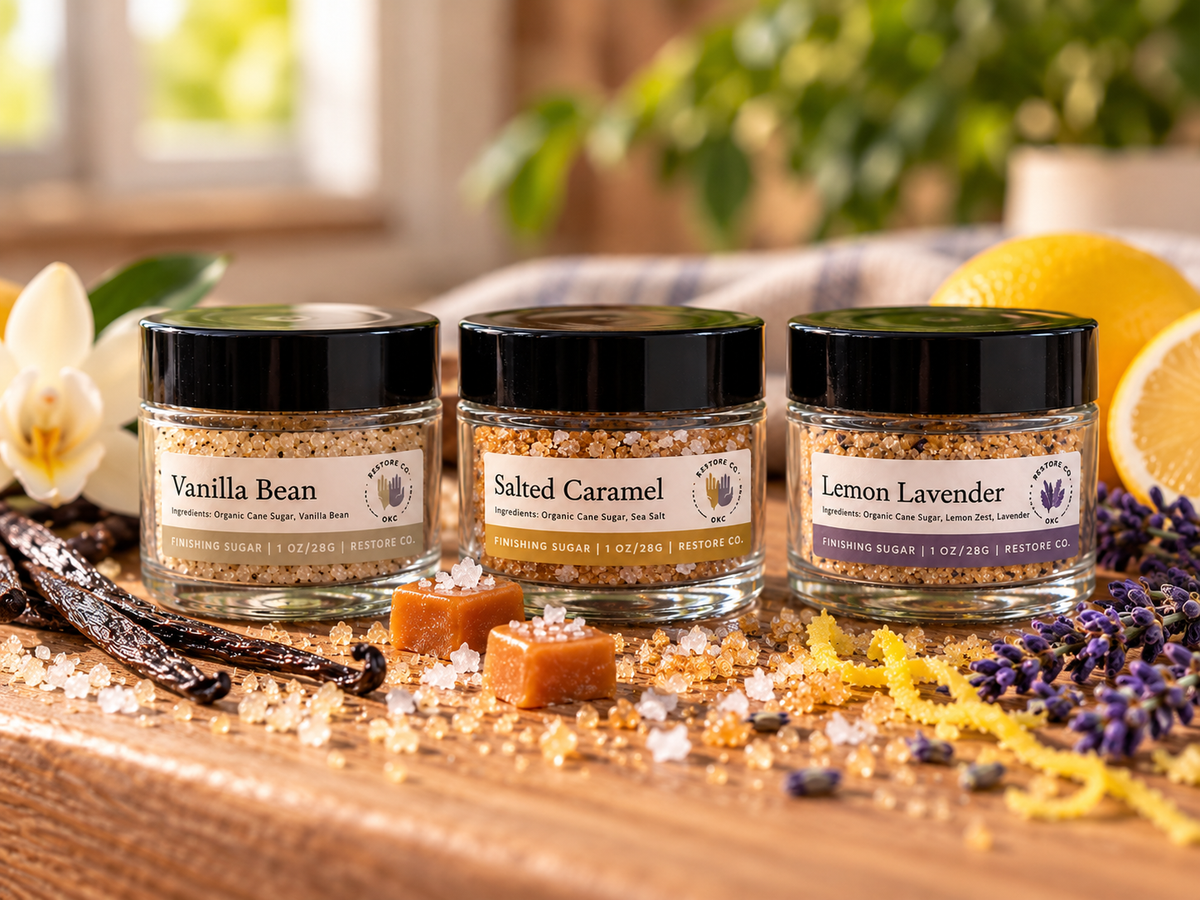 Three small glass jars of seasoning blends labeled Vanilla Bean, Salted Caramel, and Lemon Lavender, arranged on a wooden surface with spices, lemons, and flowers in a warm kitchen-like setting.