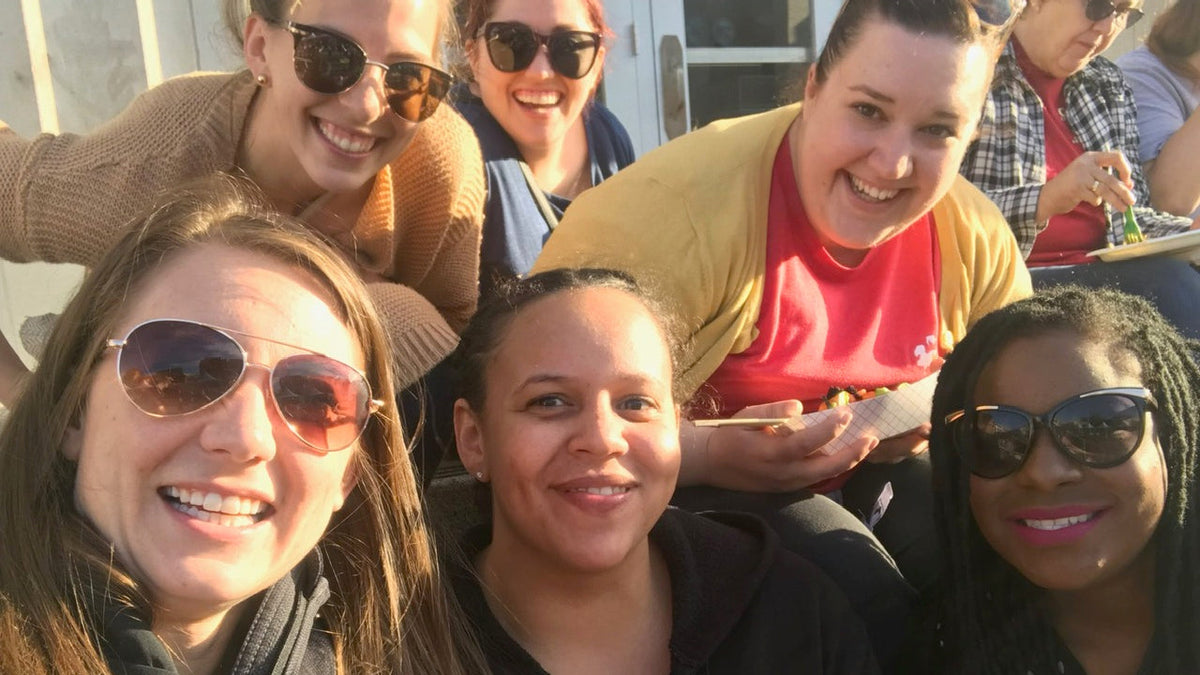 A group of five women smiling and posing together outdoors, with four of them wearing sunglasses. The photo is taken in bright natural sunlight, creating strong highlights and shadows.