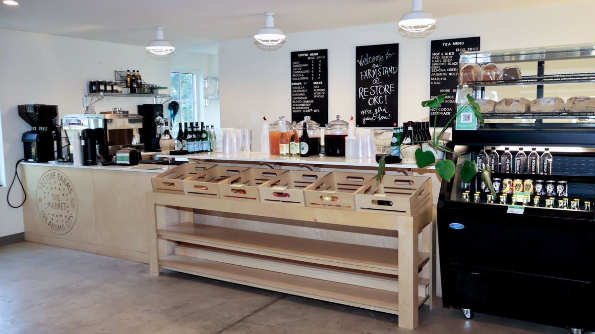 A bright, modern cafe interior featuring a light wood display table in the foreground, a wooden counter with coffee equipment, chalkboard menus on the wall, and a black beverage display case on the right.
