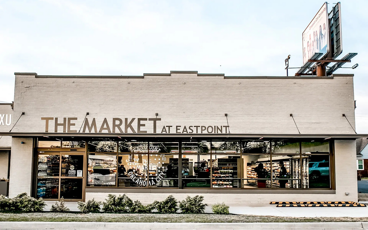The Market at Eastpoint grocery store storefront with large glass windows, a cream-colored brick facade, and a clear blue sky above.