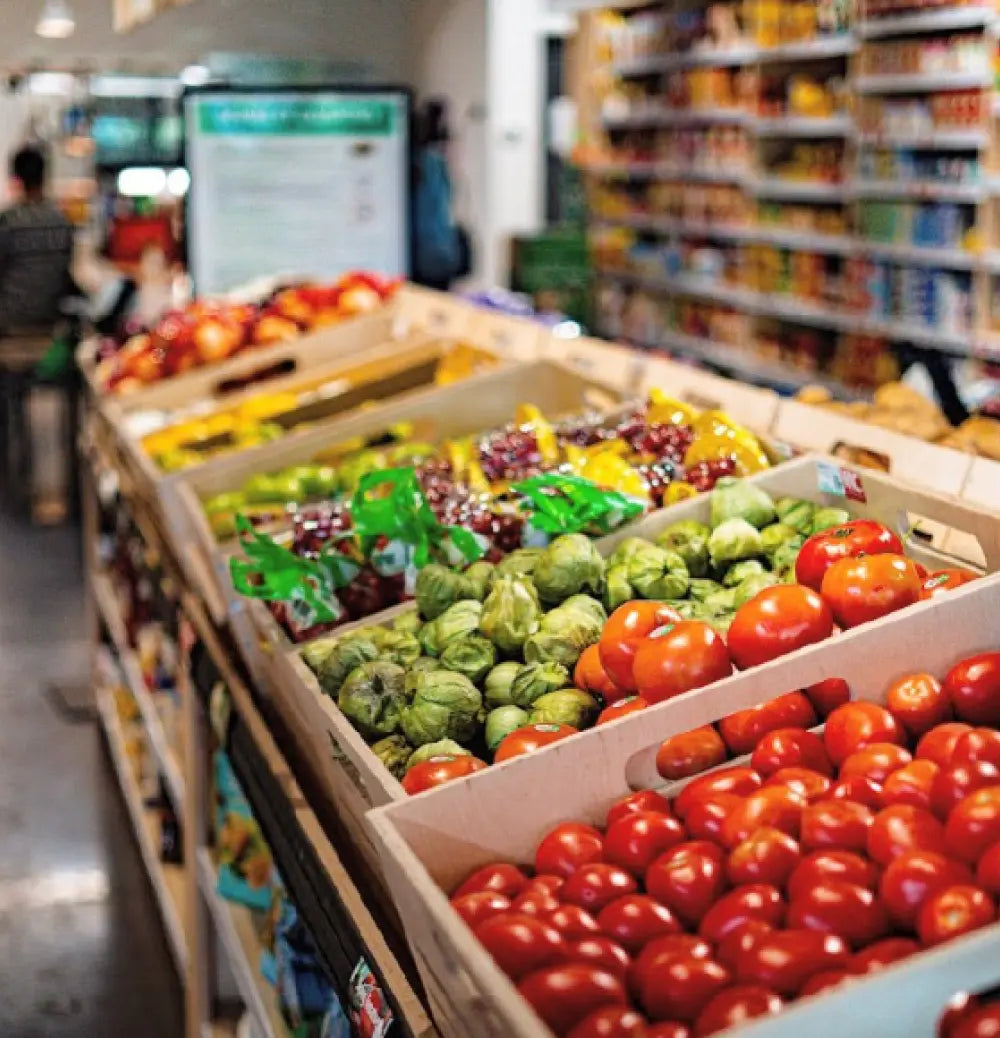 Fresh produce bins filled with red tomatoes, green Brussels sprouts, and other vegetables in a grocery store aisle.