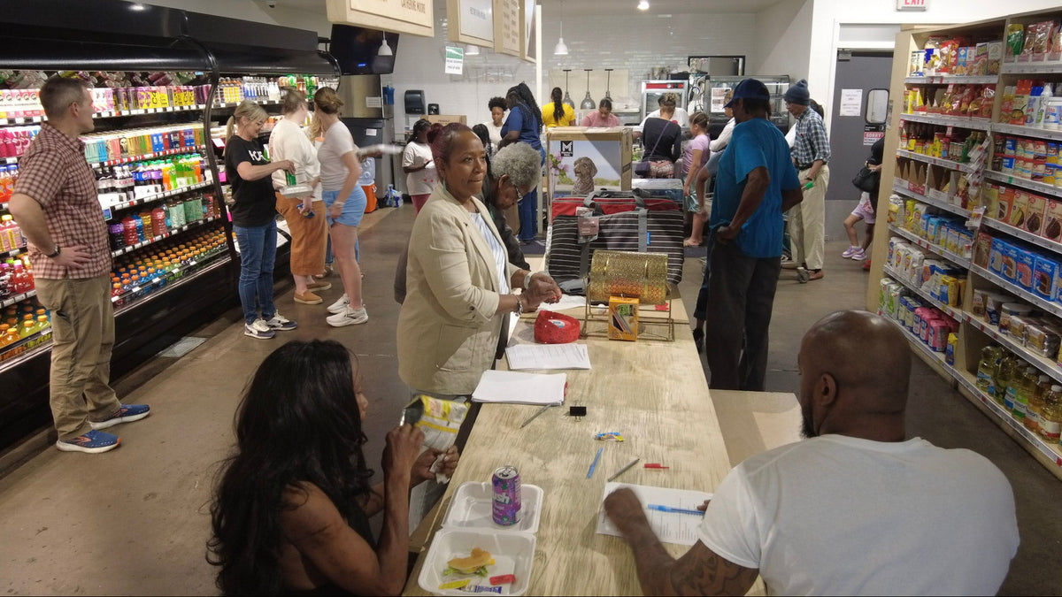 A busy grocery store interior with multiple shoppers standing at the register and aisles stocked with products on both sides. People are visible throughout the scene, so this is Lifestyle rather than Product/Packaging.